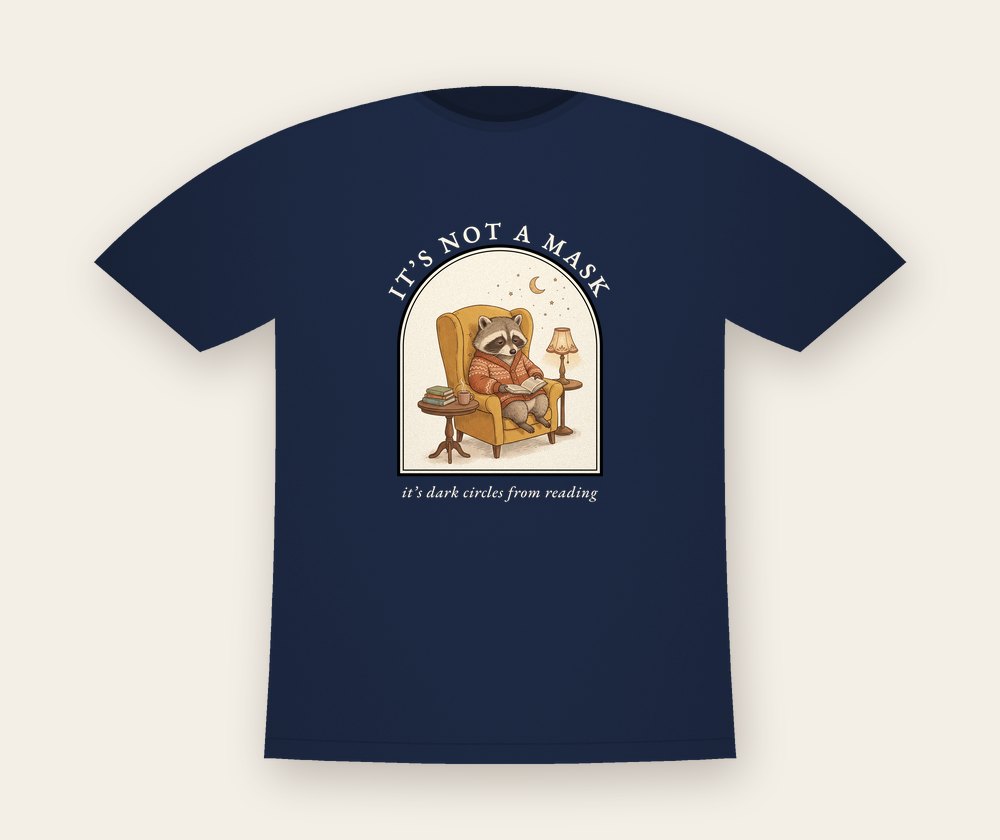 Navy T-shirt with a raccoon reading in an armchair inside an arched window, with the text It's not a mask